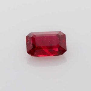 Pode incluir: Uma gema de rubi facetada, com corte retangular. A gema é de cor vermelha profunda e tem uma superfície polida. A gema está sobre um fundo branco.