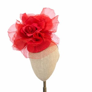 May include: A vibrant red fascinator featuring a large, layered fabric flower and delicate netting. The headpiece is mounted on a small, rounded base, creating a striking accessory for special occasions.
