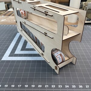 May include: A wooden storage rack with multiple shelves for holding canned goods. The rack is made of light wood and has a natural finish. The rack is designed to hold cans upright and is ideal for storing canned goods in a pantry or kitchen.