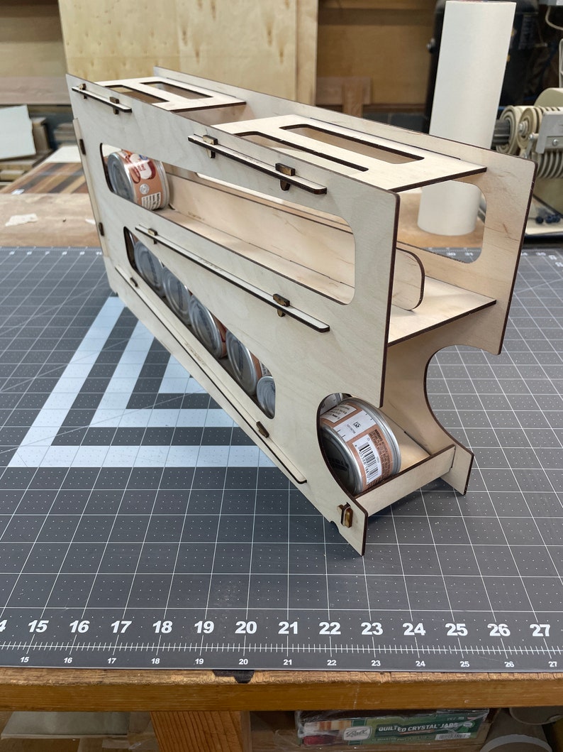 May include: A wooden storage rack with multiple shelves for holding canned goods. The rack is made of light wood and has a natural finish. The rack is designed to hold cans upright and is ideal for storing canned goods in a pantry or kitchen.
