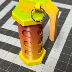 May include: A 3D-printed grenade in a gradient of yellow, orange, and red. The cylindrical body has oval cutouts, and the top features a handle and a metal ring. The design is geometric and modern.