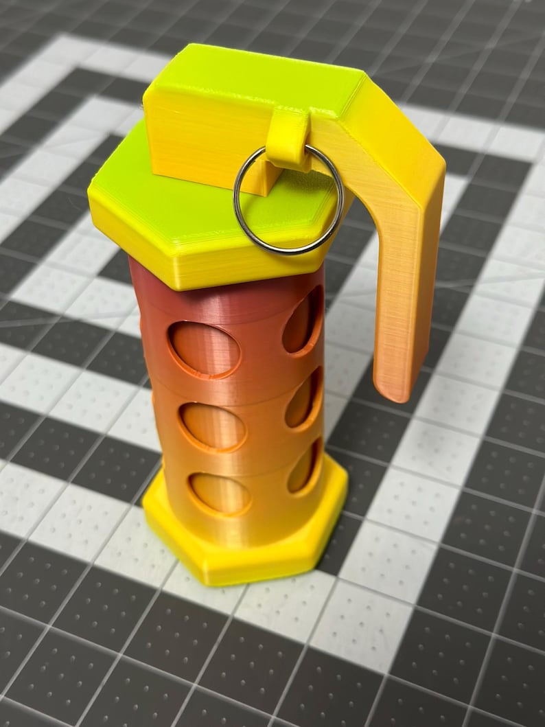 May include: A 3D-printed grenade in a gradient of yellow, orange, and red. The cylindrical body has oval cutouts, and the top features a handle and a metal ring. The design is geometric and modern.
