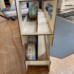 May include: A wooden storage rack with three shelves designed to hold cans of food. The rack is made of light brown wood and has a white finish. Two cans of food are visible in the rack, one on the top shelf and one on the bottom shelf.