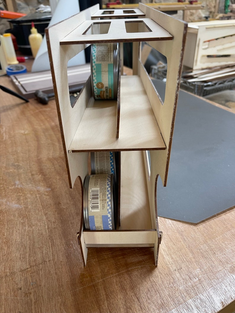 May include: A wooden storage rack with three shelves designed to hold cans of food. The rack is made of light brown wood and has a white finish. Two cans of food are visible in the rack, one on the top shelf and one on the bottom shelf.