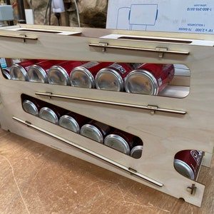 May include: A wooden can dispenser with multiple levels. The dispenser is made of light wood and has a natural finish. The dispenser is filled with red and silver cans.
