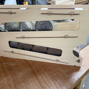 May include: A wooden storage rack with two shelves for holding canned goods. The rack is made of light wood and has a natural finish. The shelves are angled to help keep the cans from rolling out.