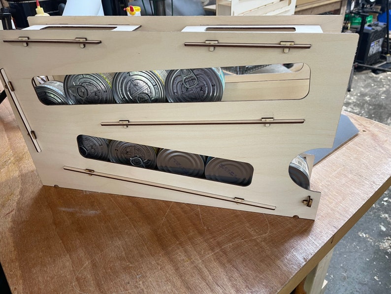 May include: A wooden storage rack with two shelves for holding canned goods. The rack is made of light wood and has a natural finish. The shelves are angled to help keep the cans from rolling out.