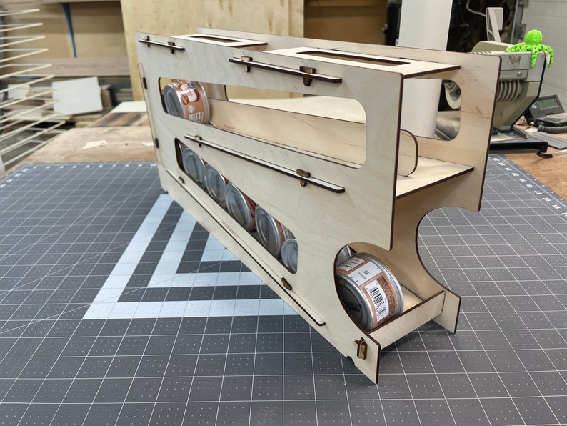 May include: A wooden storage rack for canned goods. The rack is made of light wood and has a slanted design to hold the cans in place. The rack is holding several cans of food, which are visible through the open sides of the rack.