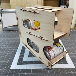 Puede incluir: Un organizador de latas de madera con tres estantes. El organizador está hecho de madera clara y tiene un acabado natural. Contiene varias latas de comida.