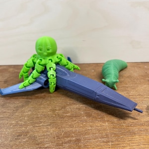 May include: A 3D-printed toy set featuring a green octopus riding a blue and gray spaceship, and a green slug. The octopus has articulated tentacles. The spaceship has blue wings. The toys are on a wooden surface.