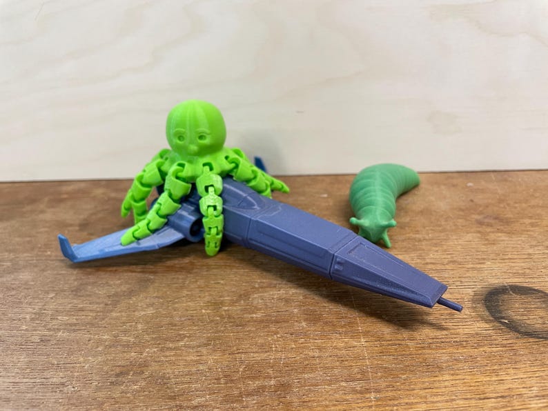 May include: A 3D-printed toy set featuring a green octopus riding a blue and gray spaceship, and a green slug. The octopus has articulated tentacles. The spaceship has blue wings. The toys are on a wooden surface.