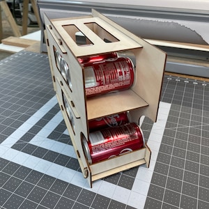 May include: A wooden can holder with three tiers, each holding four cans. The cans are red and have white labels with black text. The holder is made of light wood and has a natural finish.