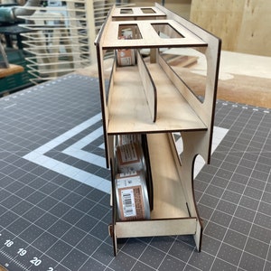 May include: A wooden storage rack with three shelves for holding cans. The rack is made of light wood and has a natural finish. The cans are visible through the open sides of the rack.