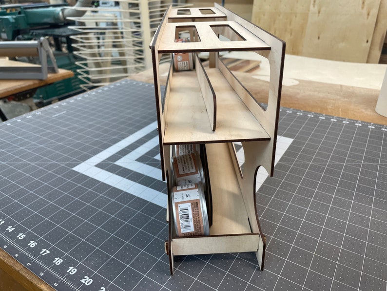 May include: A wooden storage rack with three shelves for holding cans. The rack is made of light wood and has a natural finish. The cans are visible through the open sides of the rack.