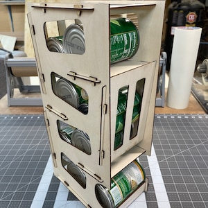 May include: A wooden can organizer with three tiers, each designed to hold multiple cans. The organizer is made of light-colored wood and has a geometric design with rectangular openings. Canned goods are visible inside the organizer.