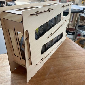 May include: A wooden storage rack with multiple slots for holding canned goods. The rack is made of light wood and has a natural finish. The rack is filled with cans of various sizes and colors.