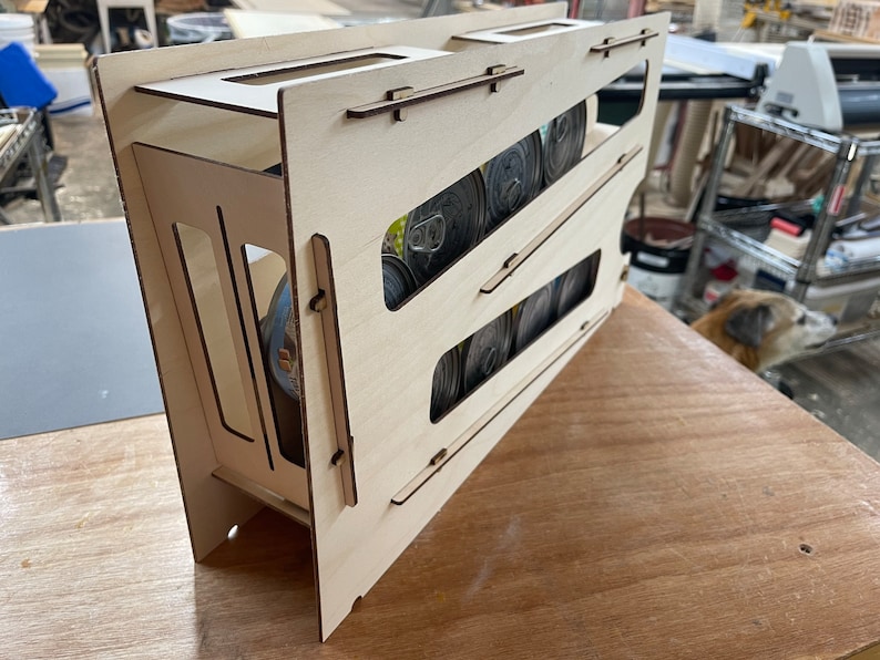 May include: A wooden storage rack with multiple slots for holding canned goods. The rack is made of light wood and has a natural finish. The rack is filled with cans of various sizes and colors.