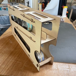 May include: A wooden storage rack with three shelves for holding canned goods. The rack is made of light-colored wood and has a sliding drawer at the bottom.