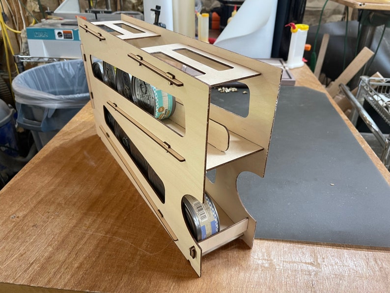 May include: A wooden storage rack with three shelves for holding canned goods. The rack is made of light-colored wood and has a sliding drawer at the bottom.