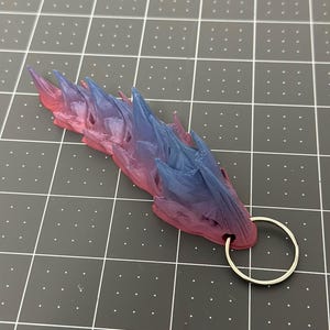 May include: A 3D-printed keychain featuring a dragon design with a gradient of pink, purple, and blue. The dragon has a series of sharp spikes along its back and a silver-colored keyring attached to its head.