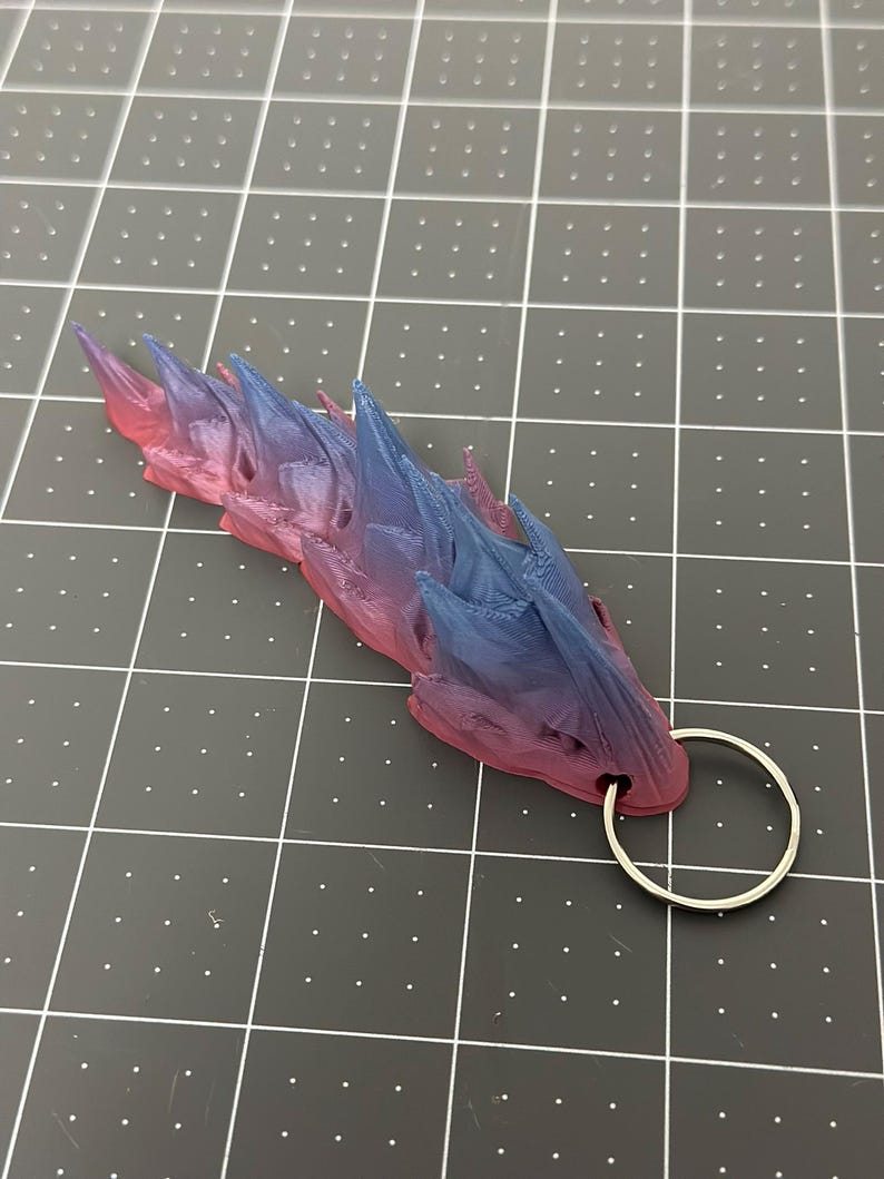 May include: A 3D-printed keychain featuring a dragon design with a gradient of pink, purple, and blue. The dragon has a series of sharp spikes along its back and a silver-colored keyring attached to its head.