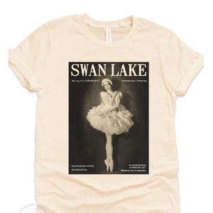 May include: Cream-colored t-shirt featuring a vintage poster design for "Swan Lake." The poster shows a ballerina in a tutu. The text "Swan Lake" is at the top in bold, black letters.