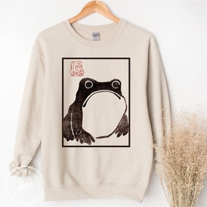 May include: A beige sweatshirt with a black and white illustration of a frog in a square frame. The frame has a red stamp with Japanese characters in the top left corner.