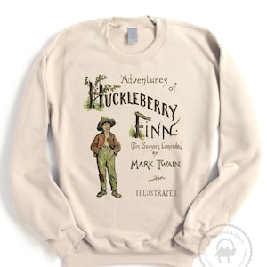 Op de afbeelding: Een beige sweatshirt met een vintage-stijl illustratie van de cover van "De avonturen van Huckleberry Finn" door Mark Twain. De illustratie toont een jonge jongen in een bruin jack en broek die voor een hek staat.