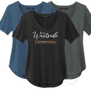 May include: Three scoop-neck t-shirts in shades of blue, black, and gray. The black shirt features the text "Westside Elementary" in white script and gold block letters. The shirts have short sleeves and a curved hemline.