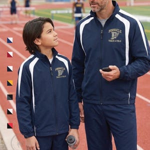 May include: Two navy blue track jackets with white accents and the text "Pinnacle Track & Field". The jackets are zipped up and worn by a man and a child on a track field. The jackets have a sporty design.
