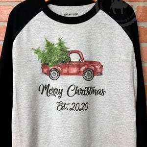 May include: A raglan-style shirt with a gray body and black sleeves. The shirt features a red truck carrying a Christmas tree, with the text "Merry Christmas Est. 2020" printed below the truck.