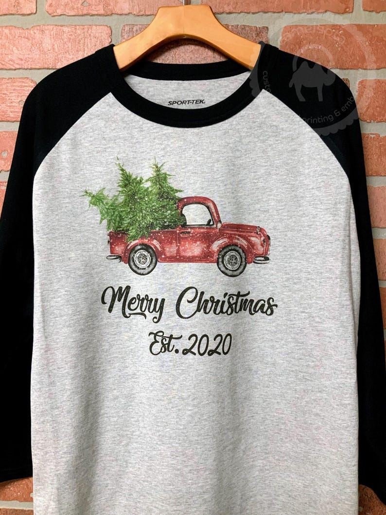 May include: A raglan-style shirt with a gray body and black sleeves. The shirt features a red truck carrying a Christmas tree, with the text "Merry Christmas Est. 2020" printed below the truck.
