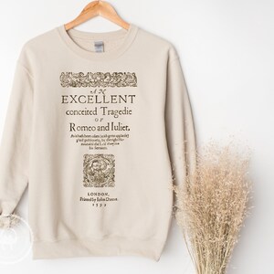 May include: A beige sweatshirt with a vintage-style print of the title page of "Romeo and Juliet" by William Shakespeare. The text reads "An Excellent Conceited Tragedie of Romeo and Juliet" and includes the publication date of 1597.