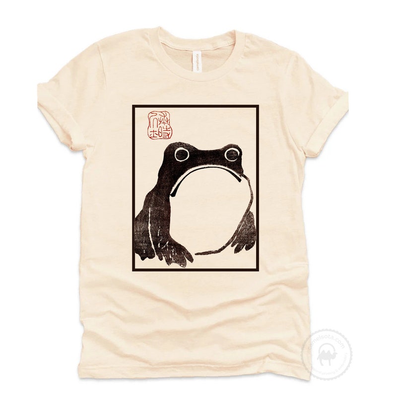 May include: A beige t-shirt with a black and white image of a frog in a square frame. The frog is looking to the left. There is a red stamp above the frog with Japanese characters.