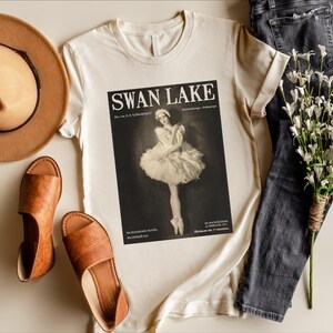 May include: Cream-colored t-shirt featuring a black and white image of a ballerina from Swan Lake. The shirt is styled with brown sandals, a tan hat, and dark wash jeans. A bouquet of white flowers completes the ensemble.