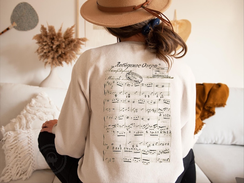 May include: Cream-colored sweatshirt with a musical score printed on the back. The text "Лебединое Озеро" is printed above the score. The sweatshirt is a casual garment, suitable for everyday wear.