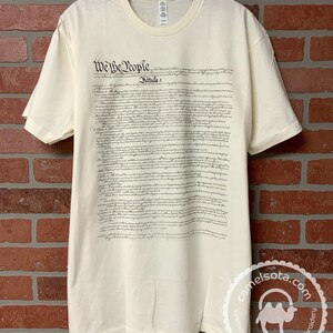 May include: A cream-colored t-shirt with the full text of the US Constitution printed on the front. The text is in black ink and includes the words "We the People".