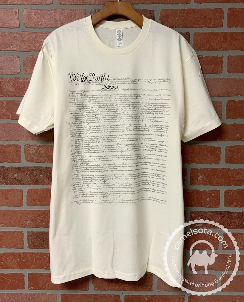 May include: A cream-colored t-shirt with the full text of the US Constitution printed on the front. The text is in black ink and includes the words "We the People".