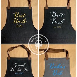 K&ouml;nnte beinhalten: Vier Sch&uuml;rzen aus Denim mit verschiedenen gestickten Texten. Die Sch&uuml;rzen sind dunkelgrau mit braunen Tr&auml;gern. Der Text auf den Sch&uuml;rzen lautet "Best Uncle Ever", "Best Dad est. 1990", "Grand Fa La La est. 2020" und "Baba's Bib".