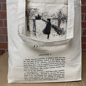 May include: A large, natural-colored canvas tote bag with a black and white illustration of a girl walking in a forest. The bag has black handles and the text "Jane" and "Chapter I." printed on the front.  There was no possibility of taking a walk that day. We had been wandering, indeed, in the leafless shrubbery an hour in the morning; but since dinner (Mrs. Reed, when there was no company, dined early) the cold winter wind had brought with it clouds so sombre and a rain so penetrating that further out-door exercise was now out of the question.