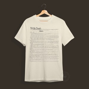 May include: A white t-shirt with the text "We the People" and "Article 1" printed on the front. The text is in a black, cursive font and is a replica of the US Constitution.