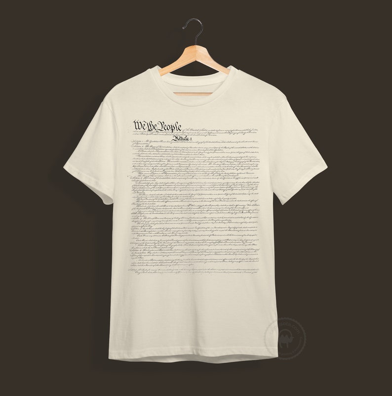 May include: A white t-shirt with the text "We the People" and "Article 1" printed on the front. The text is in a black, cursive font and is a replica of the US Constitution.