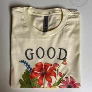 K&ouml;nnte beinhalten: Ein cremefarbenes T-Shirt mit dem Wort "GOOD" in Schwarz bedruckt. Das Shirt hat ein florales Design mit roten, gelben, rosa und blauen Blumen.