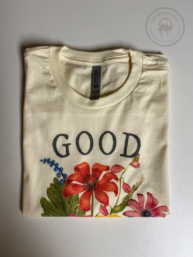 K&ouml;nnte beinhalten: Ein cremefarbenes T-Shirt mit dem Wort "GOOD" in Schwarz bedruckt. Das Shirt hat ein florales Design mit roten, gelben, rosa und blauen Blumen.