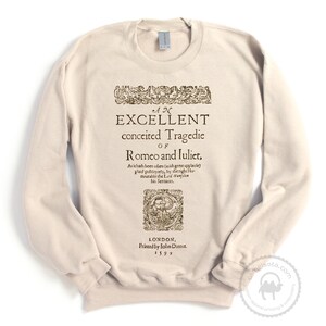 May include: A beige sweatshirt with a vintage-style print of the title page of "Romeo and Juliet" by William Shakespeare. The text reads "An Excellent Conceited Tragedie of Romeo and Juliet." The print also includes the date "1599" and the name of the printer, "John Danter."