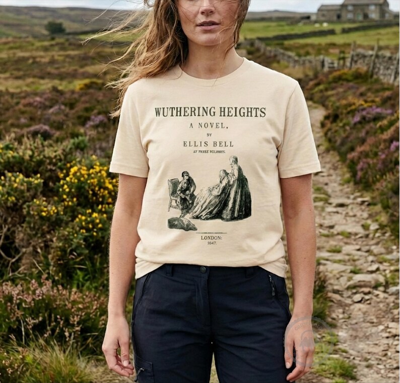 May include: Beige t-shirt featuring the text "Wuthering Heights" and an illustration from the novel. The shirt has short sleeves and is paired with dark blue pants. The image is taken outdoors.
