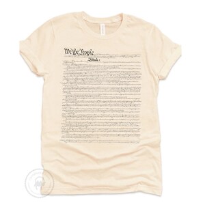 May include: A beige t-shirt with the full text of the US Constitution printed on the front. The text is in black and white and includes the words "We the People".