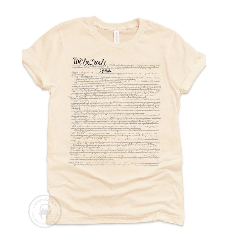 May include: A beige t-shirt with the full text of the US Constitution printed on the front. The text is in black and white and includes the words "We the People".