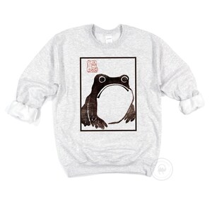 May include: A grey sweatshirt with a black and white image of a frog in a square frame. The frog is looking up and has a large, round belly. There is a red stamp in the top left corner of the frame.