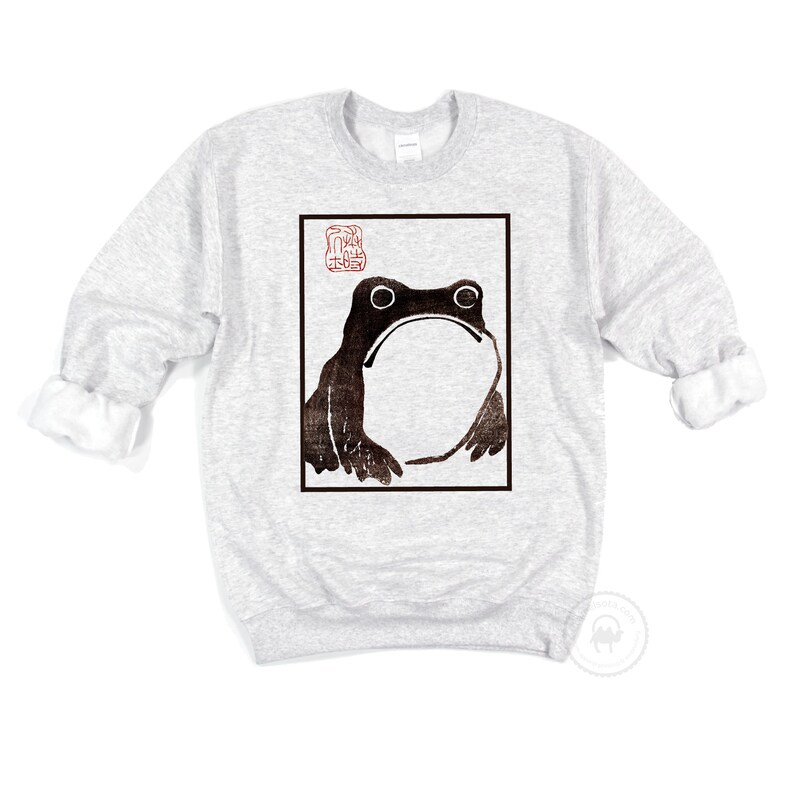 May include: A grey sweatshirt with a black and white image of a frog in a square frame. The frog is looking up and has a large, round belly. There is a red stamp in the top left corner of the frame.
